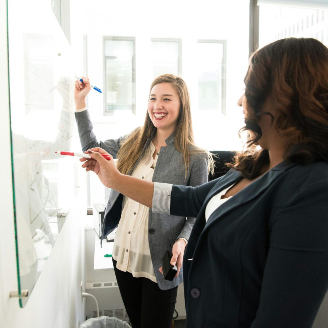 Two professional women discussing ideas on a whiteboard in a modern office setting.